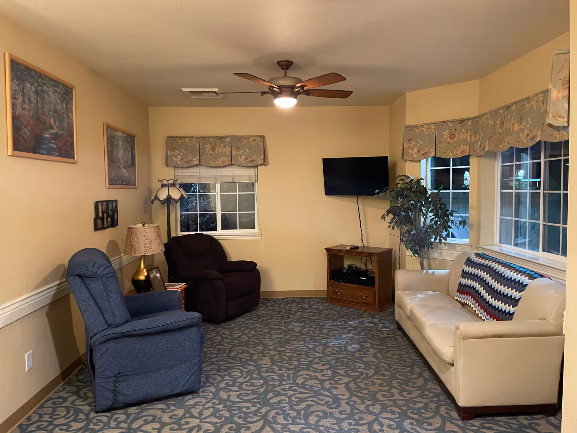 A cozy living room with a beige couch adorned with a colorful crocheted blanket, a blue recliner chair, and a dark brown armchair. The room has a patterned carpet, two windows with floral valances, a ceiling fan with a light, a wooden TV stand with a flat-screen TV mounted on the wall, and framed artwork on the walls.