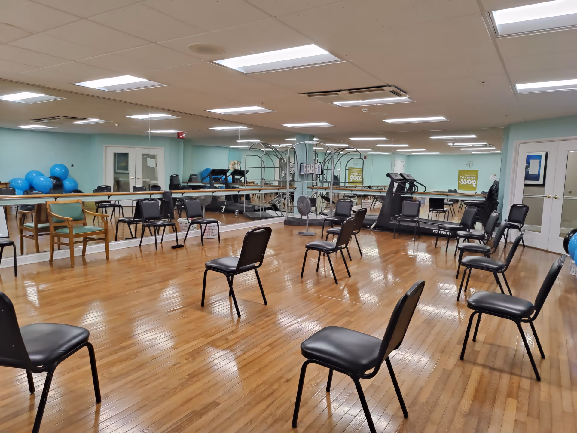 A spacious exercise room with wooden floors and light blue walls. Several black chairs are arranged in a circle in the center of the room. The room has large mirrors on one wall, exercise equipment including treadmills and blue exercise balls, and a wooden chair near the mirrors. The ceiling has recessed lighting and air conditioning vents.