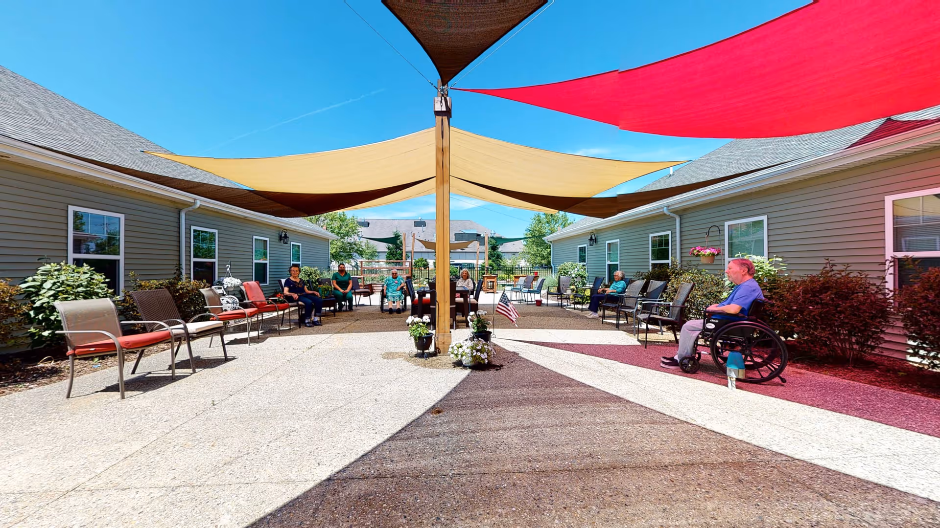 Outdoor courtyard at a retirement facility with colorful shade sails, chairs lining single-story buildings and residents seated including a person in a wheelchair.