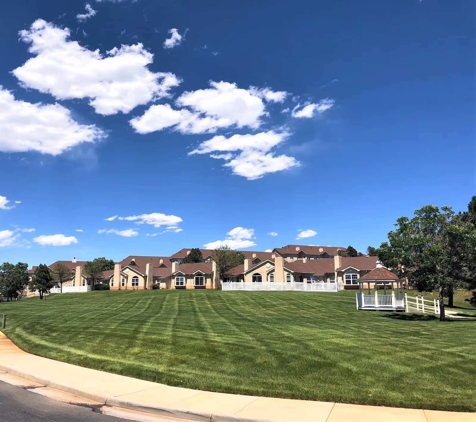 Wide view of a senior living facility named Liberty Heights with beige buildings featuring brown roofs, surrounded by a large, well-maintained green lawn under a blue sky with scattered white clouds. There is a white gazebo and some trees on the right side of the lawn.