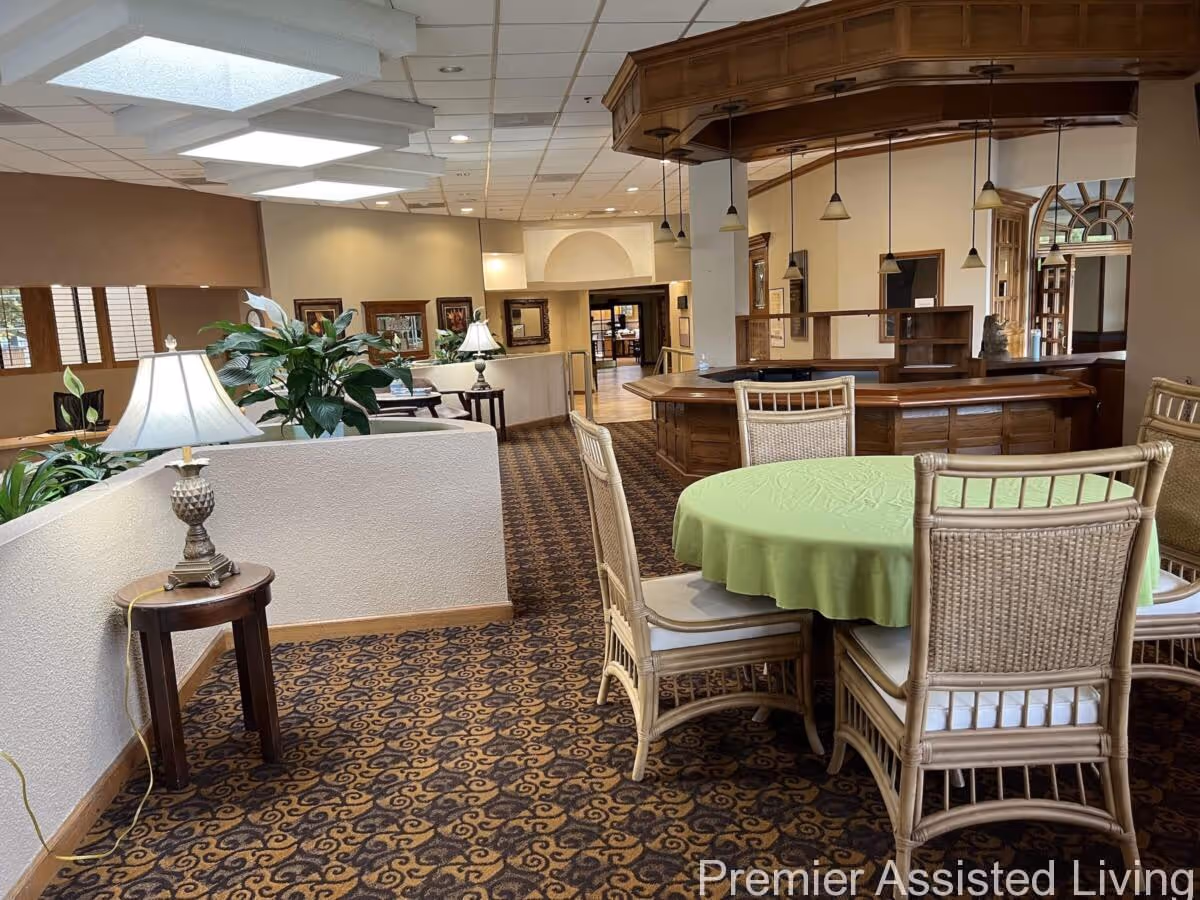 Interior view of a common area in Premier Assisted Living featuring a round table with a green tablecloth surrounded by wicker chairs, a wooden reception or bar counter with hanging pendant lights, patterned carpet, potted plants, and lamps on small tables along a white half-wall.