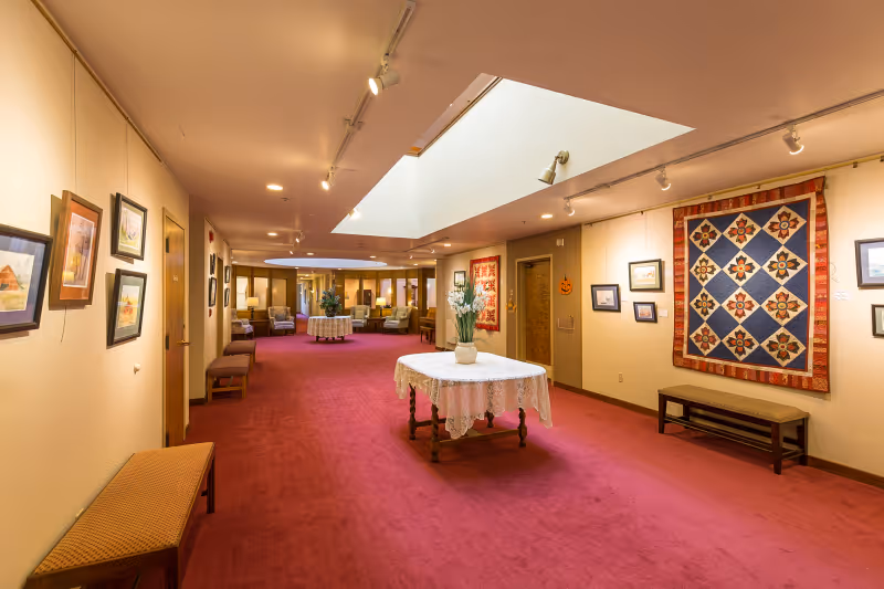 A spacious hallway in a senior living facility with red carpet flooring, beige walls adorned with framed paintings and a large quilt. There are benches along the walls and a round table with a lace tablecloth and a vase of flowers in the center. The hallway is well-lit with ceiling lights and has a skylight above.