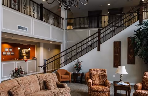 A spacious senior living facility common area with patterned upholstered sofas and armchairs, a wooden side table with a lamp, a floral arrangement on a round table, and a staircase with decorative wrought iron railing leading to an upper floor balcony.