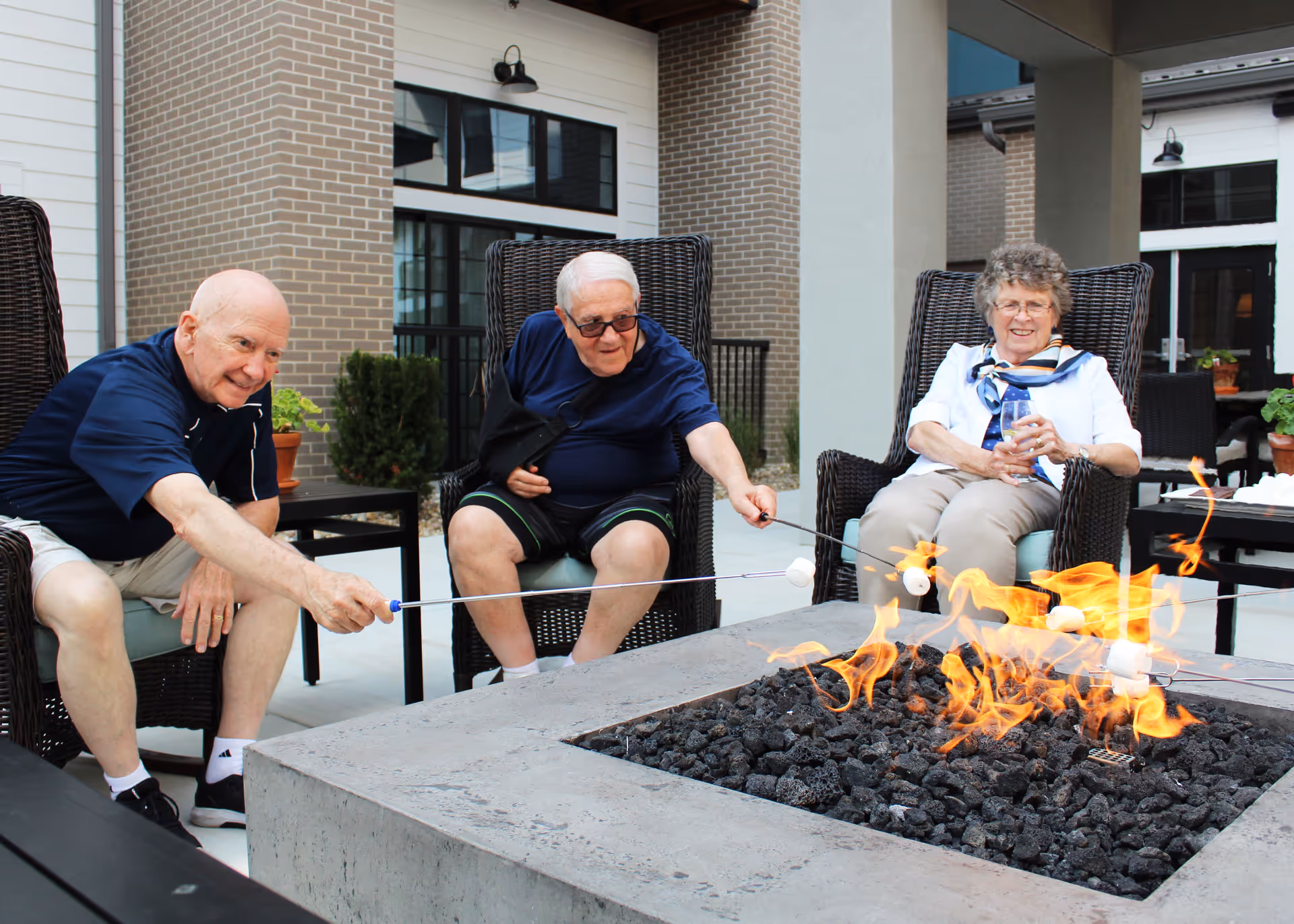 Three elderly people sitting around a modern outdoor fire pit roasting marshmallows on sticks. They are seated in wicker chairs on a patio area outside a building with brick and white siding. The atmosphere appears relaxed and social.