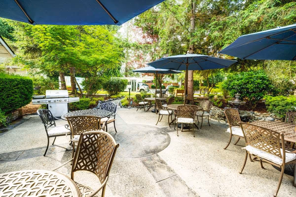 Outdoor patio area with multiple metal tables and chairs, each table shaded by large blue umbrellas. The patio is surrounded by lush green trees and bushes, with a stainless steel barbecue grill visible on the left side.