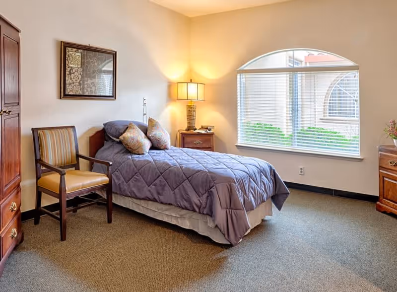A cozy bedroom with a single bed covered in a purple quilt and two patterned pillows. Next to the bed is a wooden nightstand with a lamp and a phone. A wooden chair with a yellow cushion is placed beside a wooden wardrobe. The room has a large window with blinds, allowing natural light to brighten the space. A framed artwork hangs on the wall above the bed.