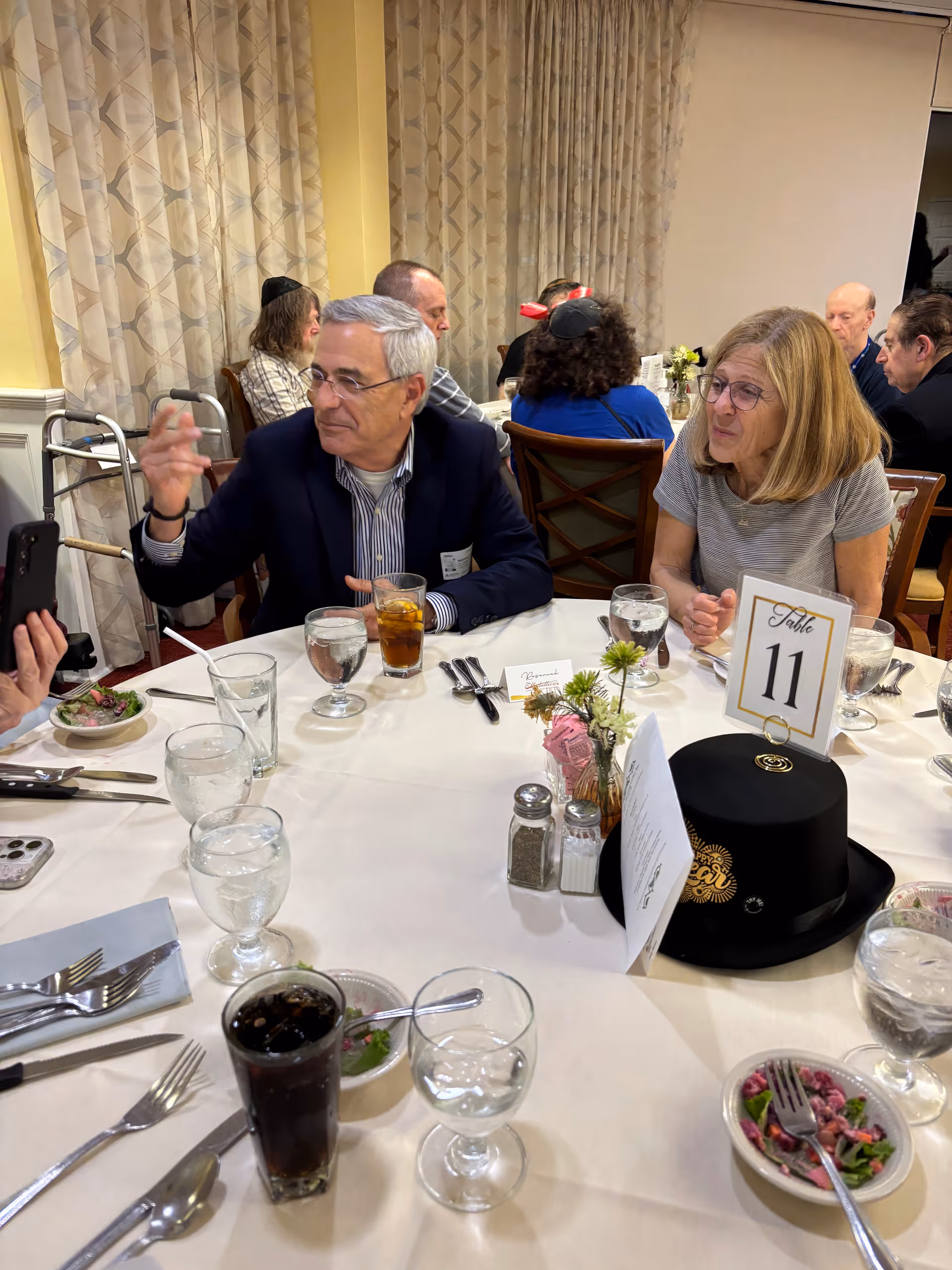 A group of older adults seated around a dining table with white tablecloth, engaged in conversation. The table is set with glasses of water, iced tea, small bowls of salad, silverware, and a black hat with a gold emblem. A table card labeled 'Table 11' is prominently displayed. The background shows curtains and more people seated at other tables.