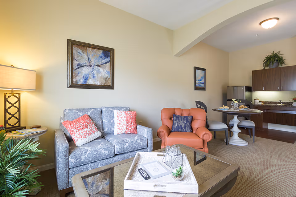 A cozy senior living room area with a patterned gray loveseat adorned with two red and white pillows, an orange armchair with a dark blue pillow, a glass coffee table with decorative items, a side table with a lamp, and a small dining area with a round table and chairs near a kitchenette with wooden cabinets and a small refrigerator.