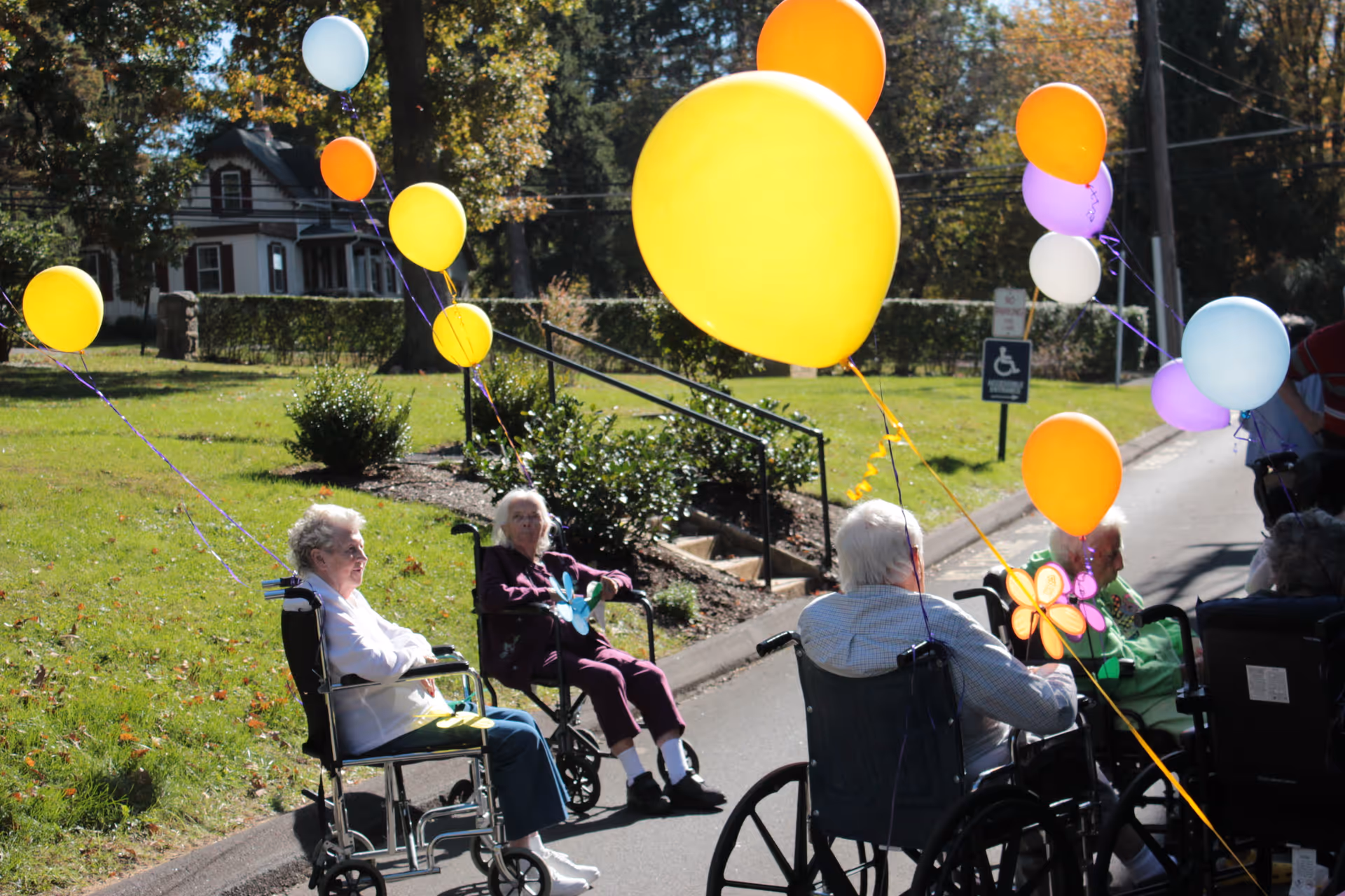 A group of elderly people sitting in wheelchairs outdoors on a sunny day, each holding colorful balloons. They are on a paved path with grass and trees in the background, and a house is visible in the distance.