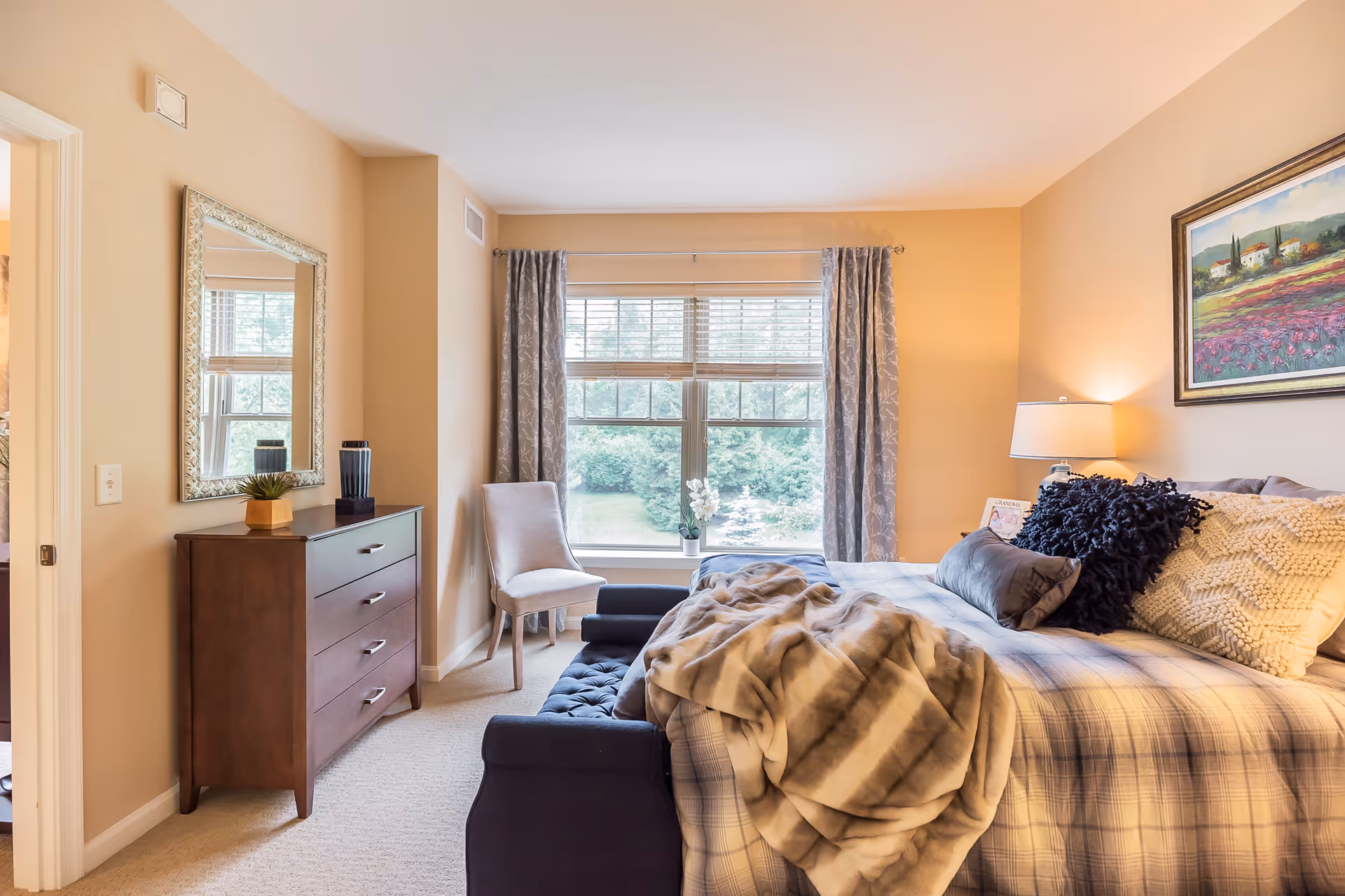 A cozy bedroom with a large window covered by gray patterned curtains. The bed is made with plaid bedding, multiple pillows, and a soft throw blanket. A dark blue tufted bench sits at the foot of the bed. To the left, there is a wooden dresser with a decorative mirror above it and a small plant and cup on top. A beige upholstered chair is positioned near the window. A framed landscape painting hangs on the wall above a bedside table with a lamp.