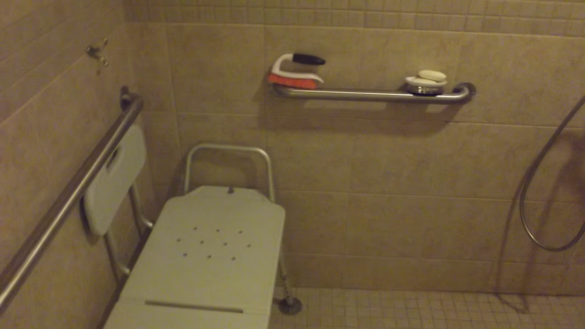 A tiled shower area with a white shower bench, grab bars on the walls, a handheld showerhead, and soap dishes holding soap and a brush.