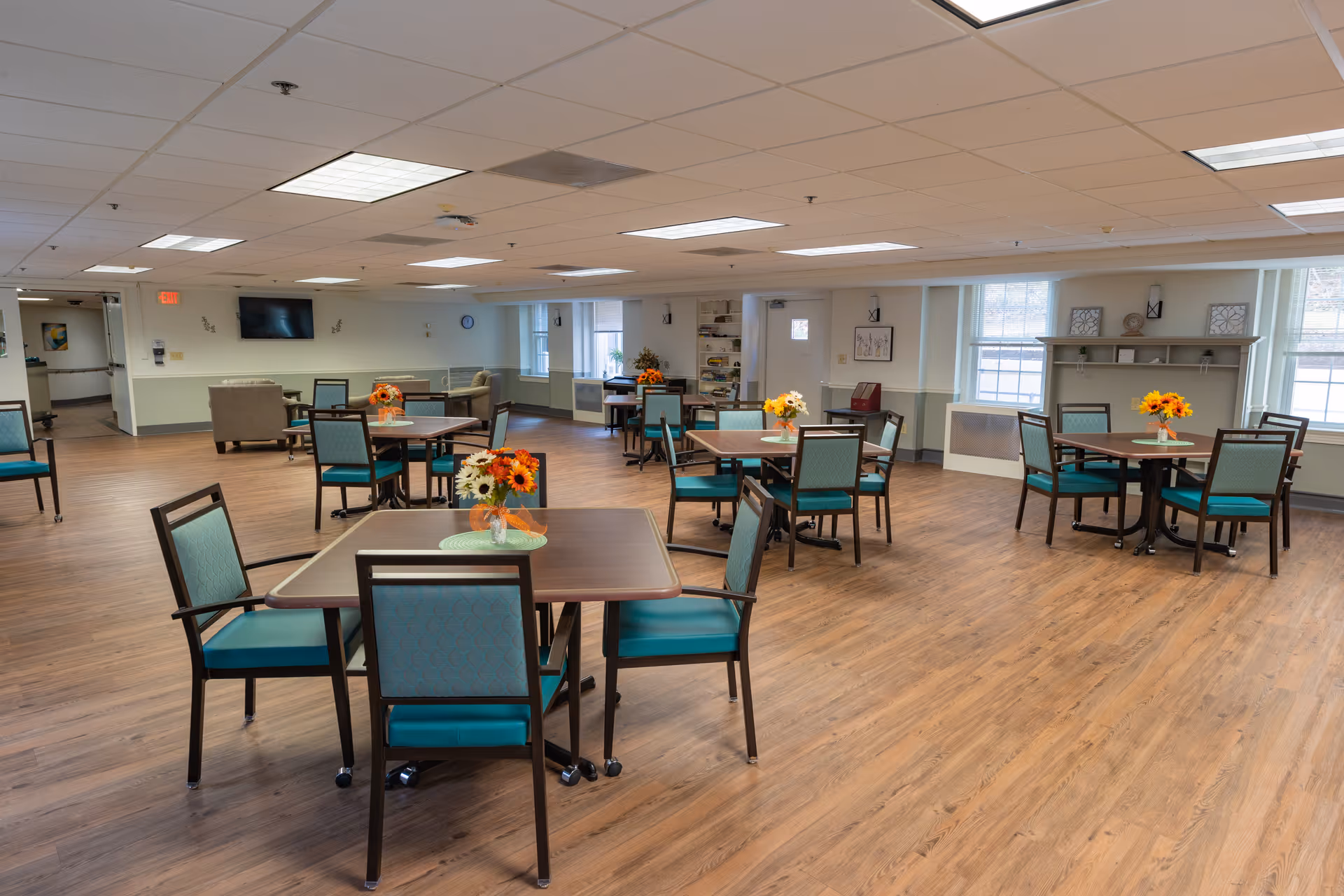 A spacious dining room with several square tables, each surrounded by four chairs with teal cushions. Each table has a small vase with colorful flowers. The room has wood flooring, white walls with a light green lower half, and large windows letting in natural light. There is a television mounted on one wall and some seating areas with armchairs in the background.