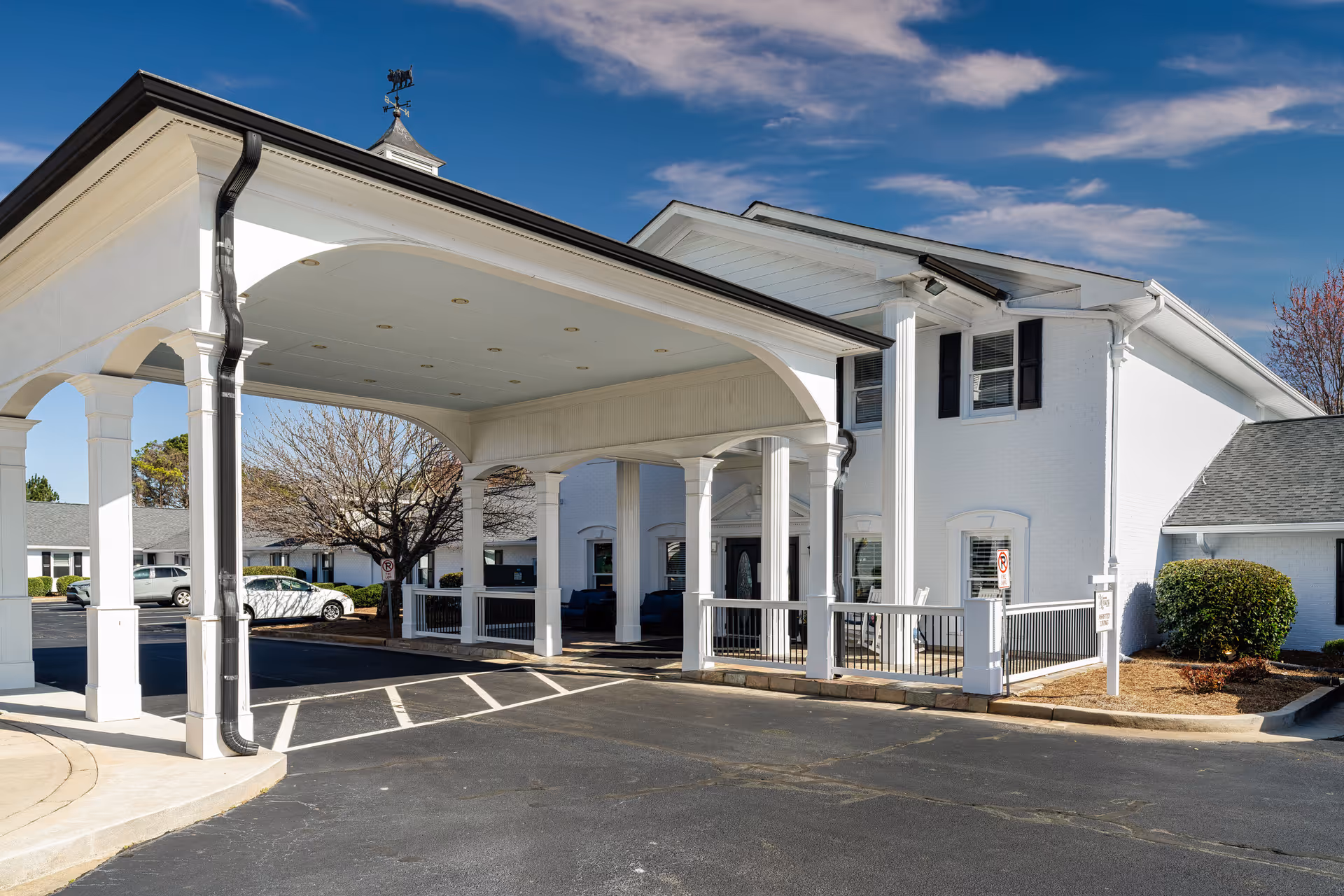 Exterior view of a white senior living facility building with a covered entrance supported by white columns. There are parked cars and some landscaping with bushes and trees around the building under a partly cloudy blue sky.