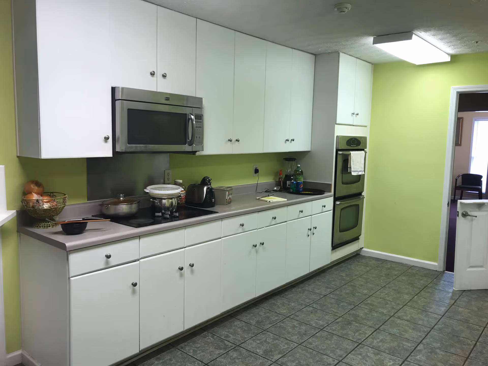 A kitchen with white cabinets and drawers, a gray countertop, and green walls. The kitchen features a built-in microwave above a stovetop, a double oven, a toaster, a blender, and various kitchen items such as a bowl of onions, a pot, and bottles. The floor is tiled with gray tiles, and there is an open door leading to another room with a chair visible.