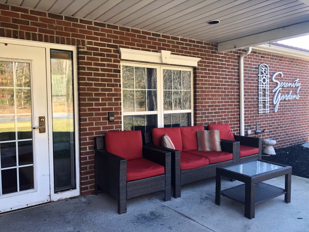 Covered brick entrance patio with wicker seating topped with red cushions and a 'Serenity Gardens' sign on the wall.