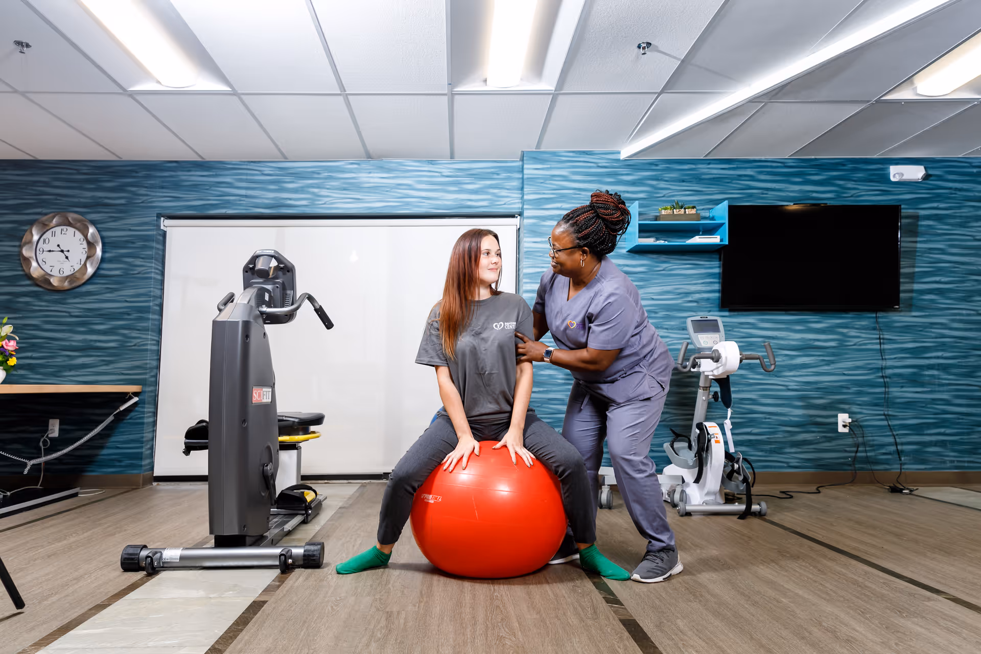 A woman in gray scrubs assists a young woman sitting on a large red exercise ball in a fitness room with exercise equipment, a wall clock, and a mounted TV in the background.