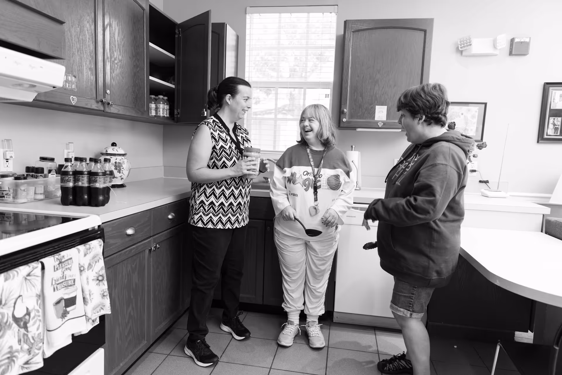 Three people standing and chatting in a kitchen area with wooden cabinets, a stove, and a window in the background. One person is holding a drink, and all appear to be smiling and enjoying the conversation.