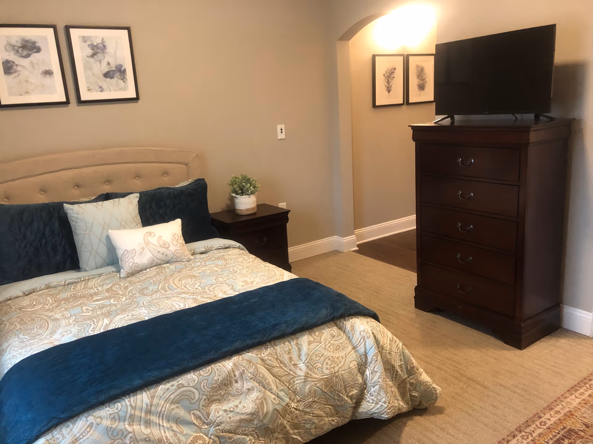 A bedroom with a beige upholstered headboard bed dressed in patterned bedding with blue and beige tones, three decorative pillows, a dark wooden nightstand with a small potted plant, and a dark wooden dresser with a flat-screen TV on top. The walls are beige with framed artwork above the bed and in the hallway.