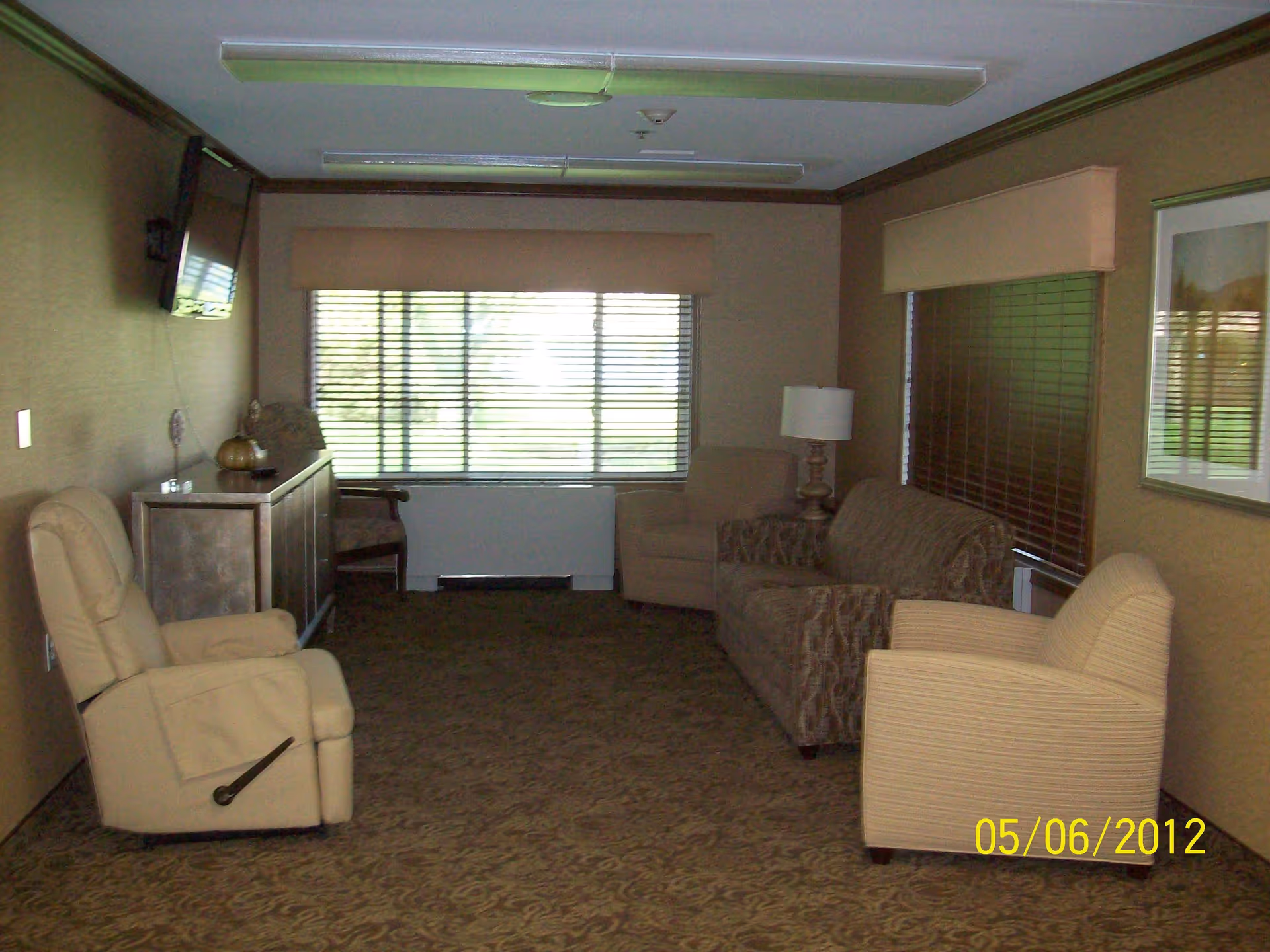 A long communal sitting room with a recliner, armchairs, a sofa, a wall-mounted TV, and windows with blinds.