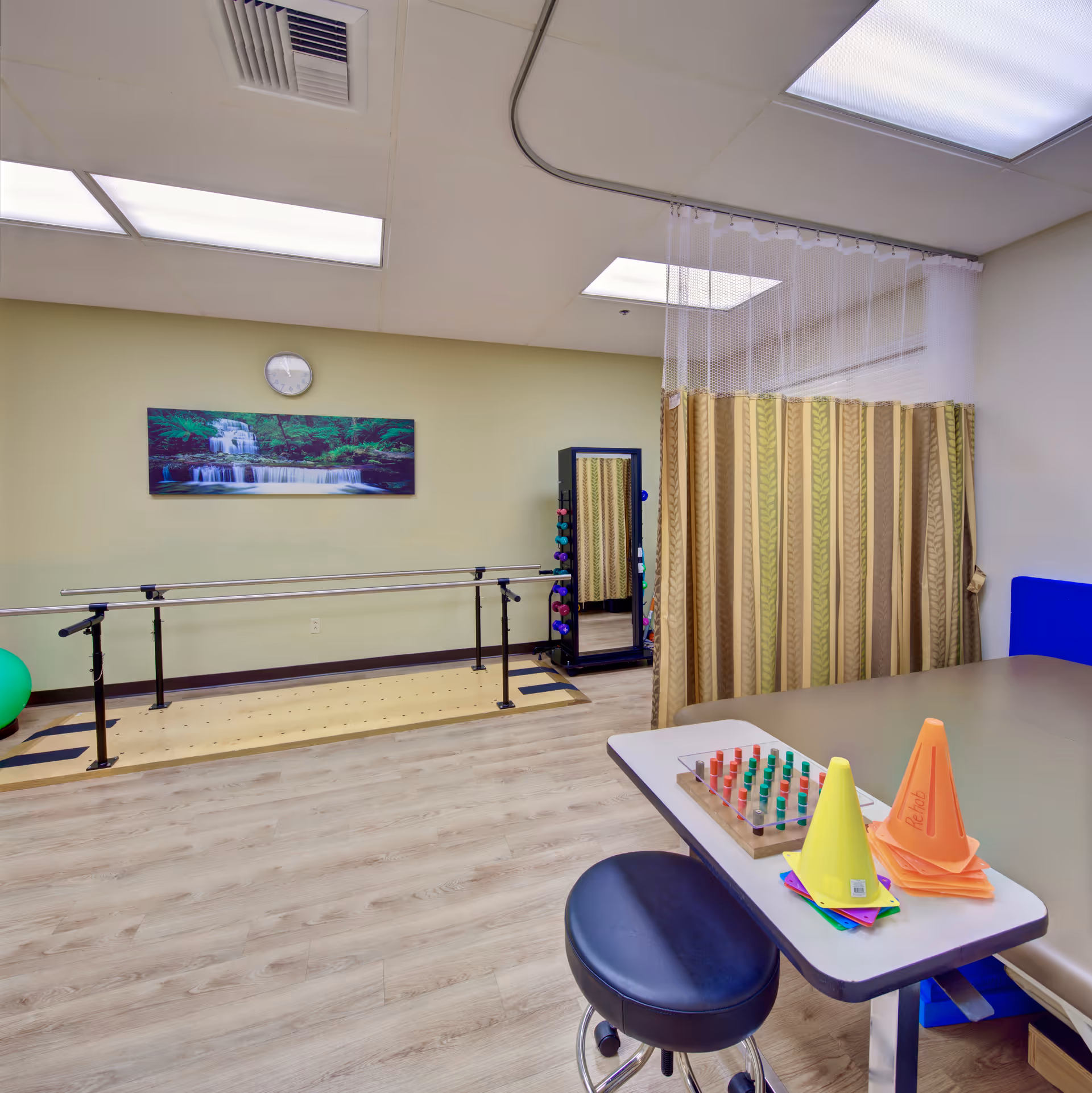 A rehabilitation room with parallel bars for walking exercises, a green exercise ball, a rack of colorful dumbbells, a wall clock, and a nature-themed painting. There is a table with colorful cones and a pegboard game, a stool, and a privacy curtain in the corner.