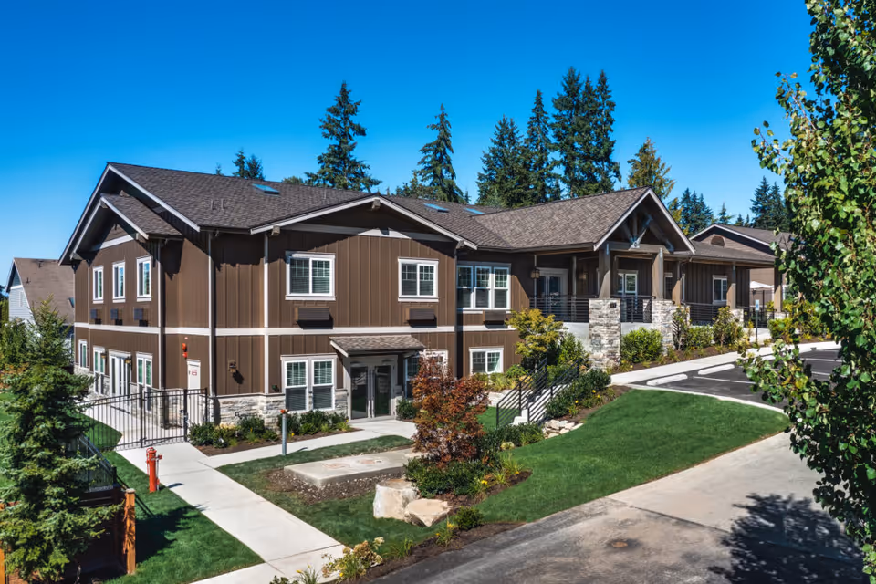 Exterior view of a two-story brown senior living facility building with white-trimmed windows, surrounded by landscaped greenery and trees under a clear blue sky.