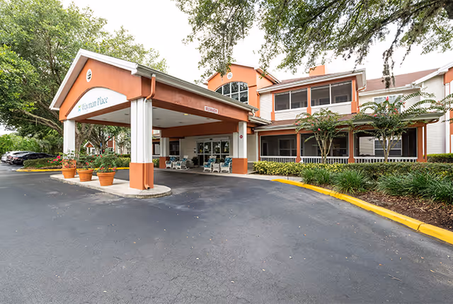 Exterior view of Wayman Place senior living facility showing the entrance with a covered drop-off area supported by white columns. There are potted plants near the entrance and a driveway with a yellow curb. The building has two stories with large windows and a porch area with seating. Trees and landscaping surround the building.