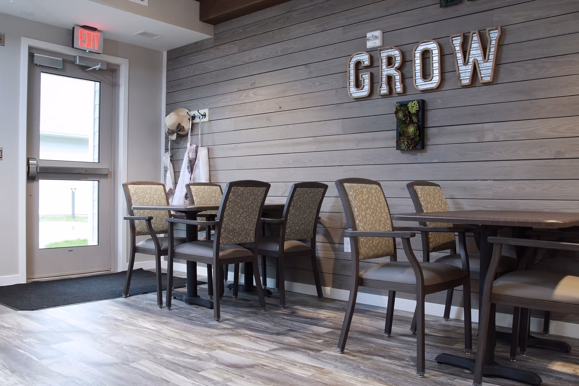 Interior view of a dining area with several tables and chairs arranged neatly. The wall is decorated with large letters spelling 'GROW' and a small vertical planter with green plants. There is a door with a window and an exit sign above it on the left side.