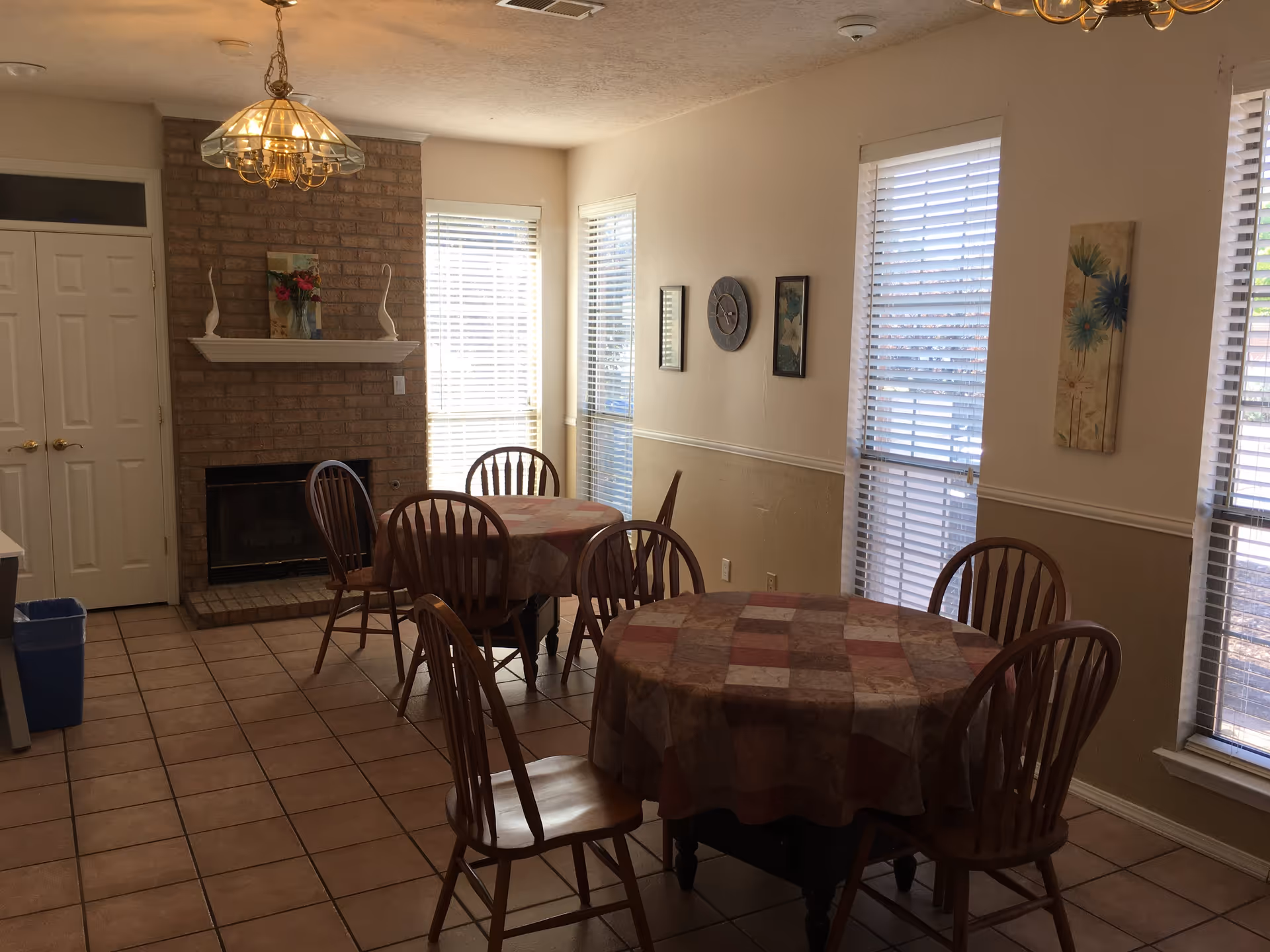 Interior view of a dining area with two round tables covered with patterned tablecloths, each surrounded by wooden chairs. The room has tiled flooring, a brick fireplace with decorative items on the mantel, and several windows with blinds letting in natural light. Wall decorations and a hanging light fixture are also visible.