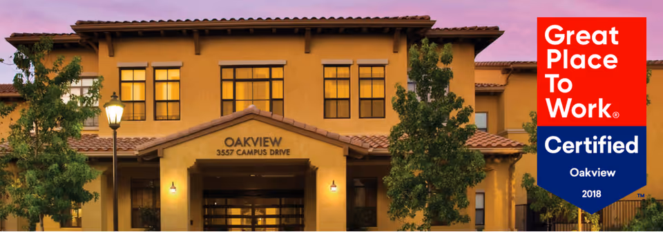 Front exterior view of OakView Health Center building at dusk with warm lighting, trees, and a street lamp in front. A red and blue banner on the right side reads 'Great Place To Work Certified Oakview 2018'.