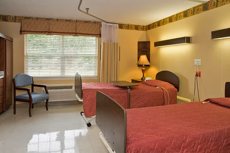 A nursing center bedroom with two hospital-style beds covered in red bedspreads. There is a wooden chair with blue upholstery near a window with blinds and a valance. A bedside table with a lamp is positioned between the beds, and a privacy curtain hangs from a ceiling track. The walls are painted a soft yellow, and the floor is tiled.