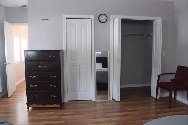 A bedroom interior with wooden flooring, a dark wooden chest of drawers on the left, two white closet doors in the center, a tall mirror leaning against the wall between the closets, a clock above the mirror, and a wooden chair with red upholstery on the right. The reflection in the mirror shows part of a bed with a white pillow and headboard.
