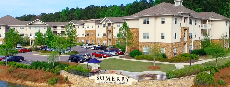 Exterior view of Somerby St. Vincent’s One Nineteen senior living facility showing a multi-story building with beige and brick facade, surrounded by green trees and a parking lot with several cars. There is a stone wall with a sign displaying the facility name in front of the building.