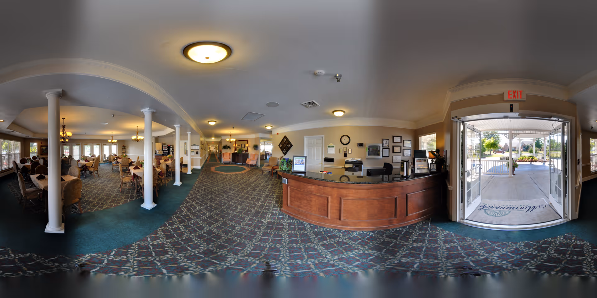 Interior view of a senior living facility lobby and dining area. The image shows a reception desk on the right side near the entrance with glass doors leading outside. To the left, there is a spacious dining room with tables and chairs, where several seniors are seated. The area is well-lit with ceiling lights and chandeliers, and the floor is covered with patterned carpet. White columns separate the dining area from the hallway leading further into the facility.