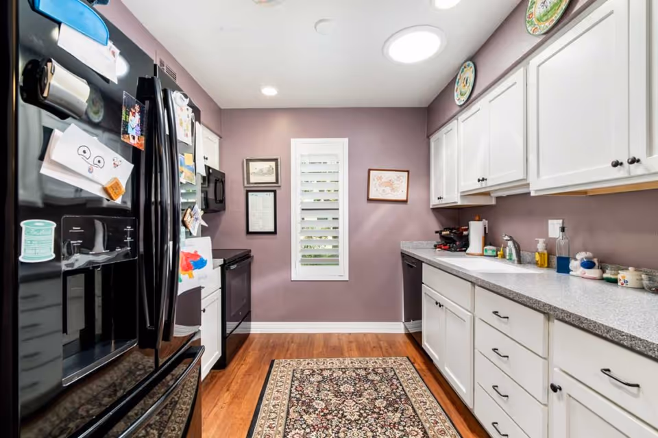 A modern kitchen with white cabinets and gray countertops. The walls are painted purple, and there is a window with white shutters at the end of the room. The kitchen features a black refrigerator with various magnets and drawings attached, a black oven, and a dishwasher. Decorative plates are displayed above the cabinets, and a patterned rug covers part of the wooden floor.