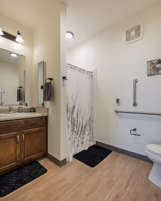 A clean and accessible bathroom with a wooden vanity and sink on the left, a large mirror above it, a shower with a white curtain featuring a plant design, a toilet on the right, grab bars on the wall, and light wood flooring.