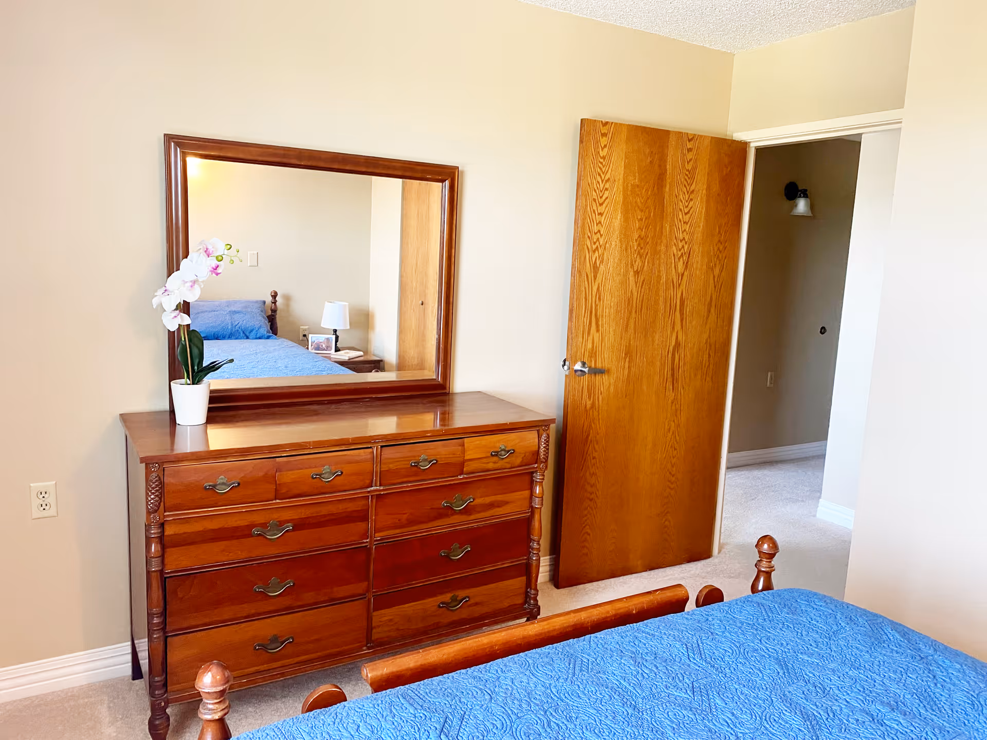 Bedroom with a wooden dresser and mirror topped by a potted orchid, a blue-covered bed, and an open wooden door.