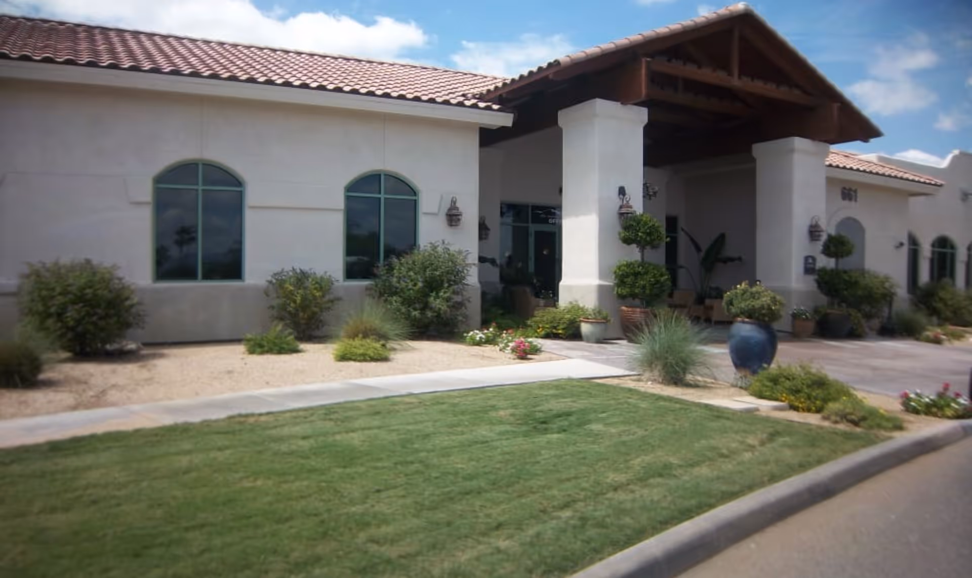 The front exterior and covered entrance of a senior living building with arched windows, potted plants, and manicured landscaping.