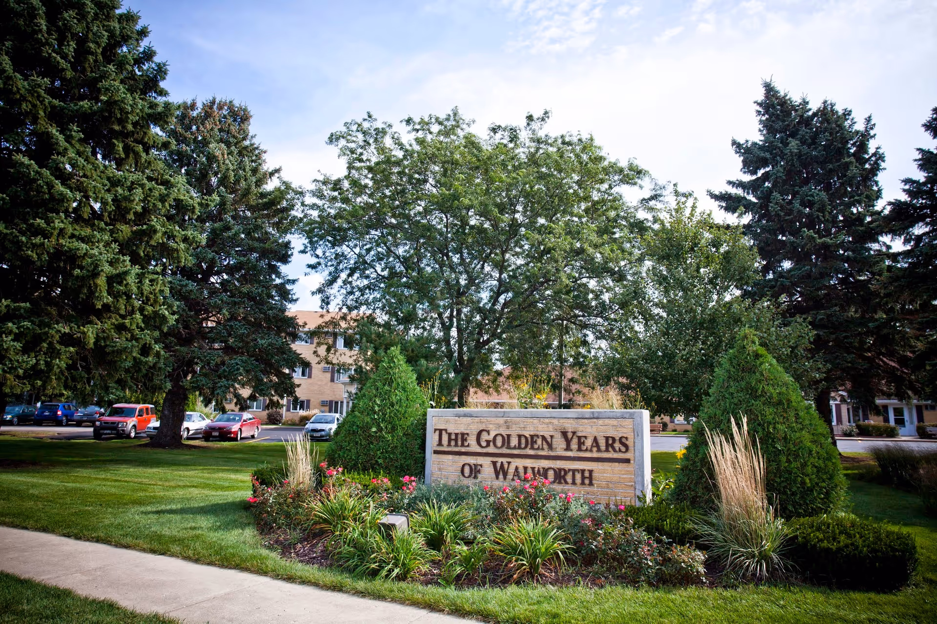Outdoor view of the entrance area to The Golden Years of Walworth senior living facility, featuring a landscaped garden with flowers, bushes, and trees surrounding a stone sign with the facility's name. In the background, there is a parking lot with several cars and a multi-story building partially visible behind the trees.