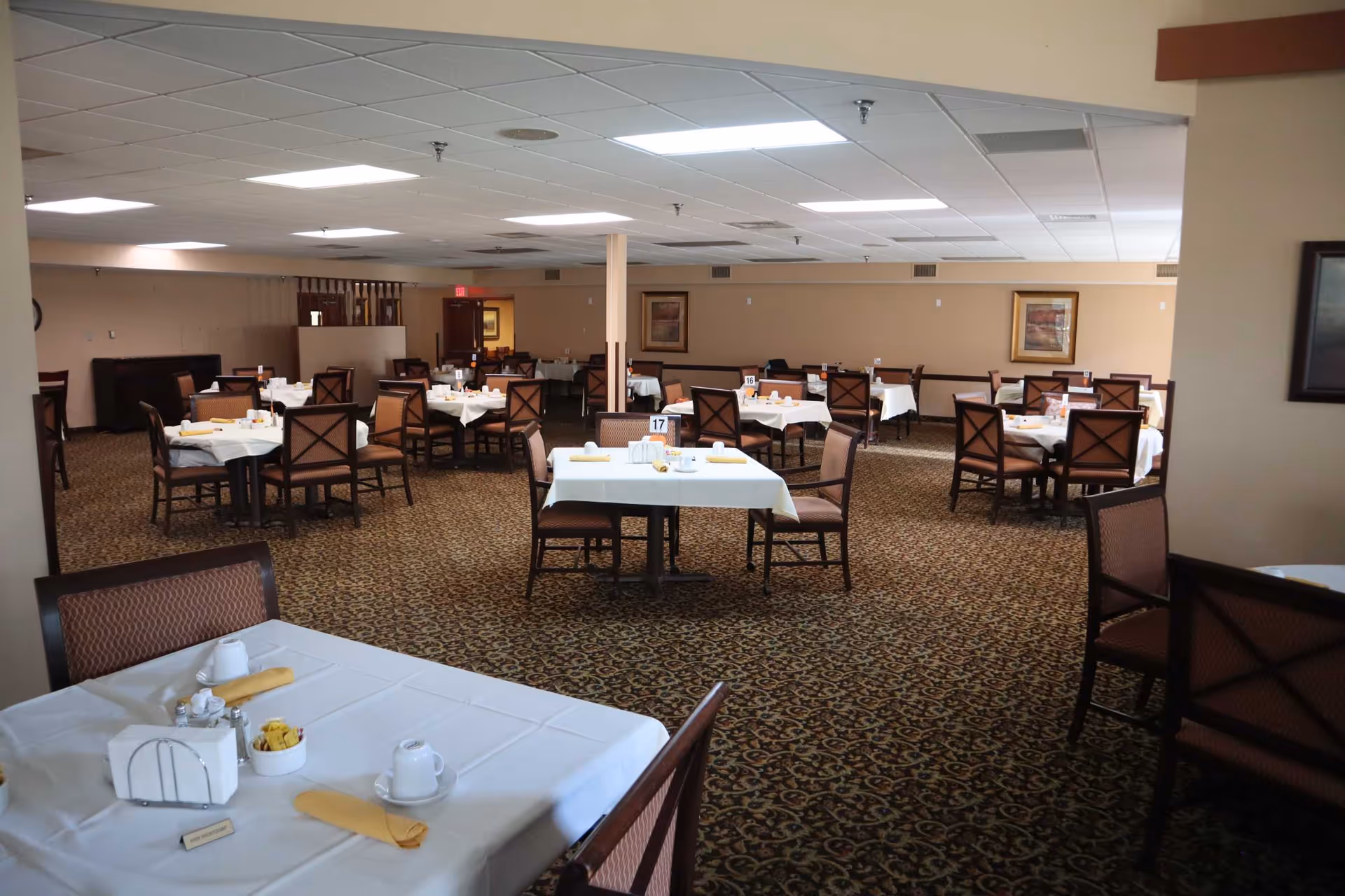 A spacious dining room with multiple tables covered in white tablecloths, each set with cups, napkins, and condiments. The room has patterned carpet flooring, beige walls with framed artwork, and a drop ceiling with fluorescent lighting. Chairs with cushioned seats and backs are arranged around the tables.