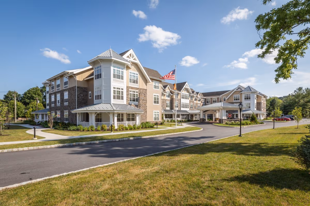 Exterior view of a large, modern senior living facility building with multiple windows, a covered entrance, and an American flag flying on a flagpole. The building is surrounded by well-maintained lawns, trees, and a paved driveway under a clear blue sky with some clouds.