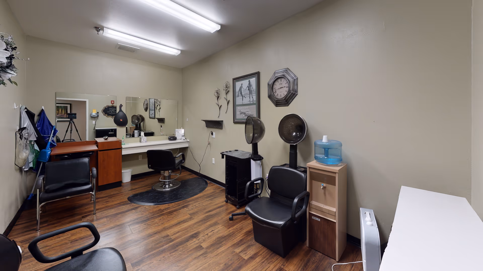 Interior view of a senior living facility's hair salon room with wooden flooring, beige walls, two black salon chairs, two hair dryers, a large mirror, a clock, framed artwork, and a water dispenser on a small cabinet.