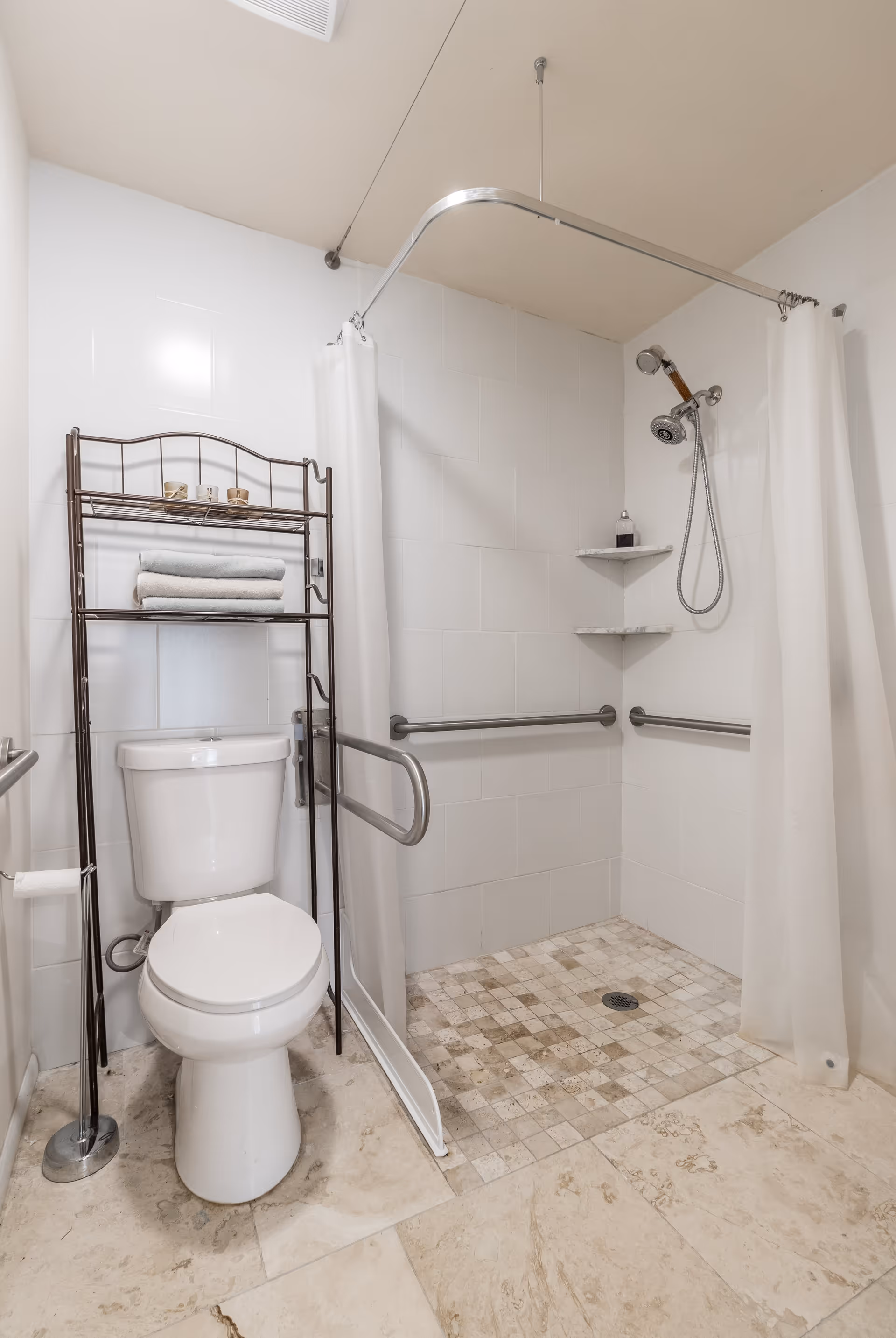 A clean bathroom with a white toilet, a metal rack holding folded towels and small containers above the toilet, and a walk-in shower with a handheld showerhead, grab bars, and a white shower curtain. The floor is tiled with beige and light brown tiles.
