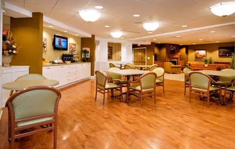 A spacious, well-lit common area with wooden flooring, several round tables surrounded by green cushioned chairs, a counter with a coffee station and a wall-mounted TV, and comfortable seating areas with sofas and armchairs in the background.