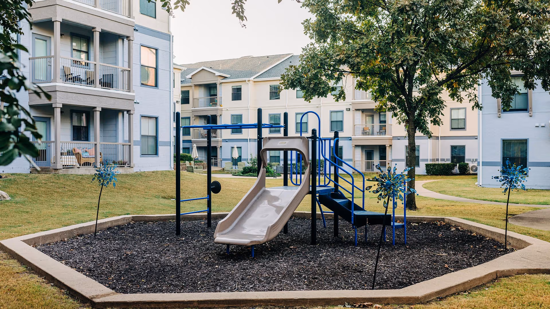 Outdoor playground area with a slide and climbing structure surrounded by a mulch bed, located in the courtyard of a senior living facility with multi-story residential buildings and trees in the background.