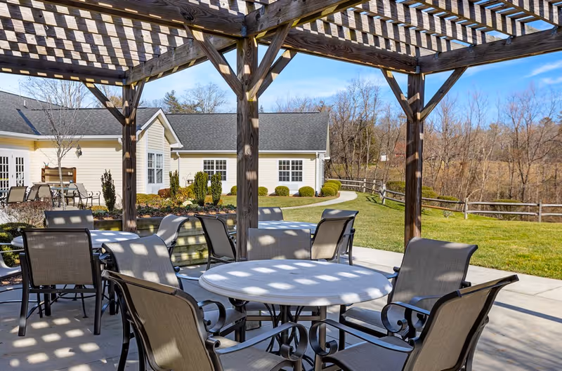 Outdoor patio area at Carillon Assisted Living of Hendersonville featuring round tables with chairs under a wooden pergola casting shadows. In the background, there is a beige building with white trim, landscaped bushes, a green lawn, and a wooden fence with leafless trees beyond.
