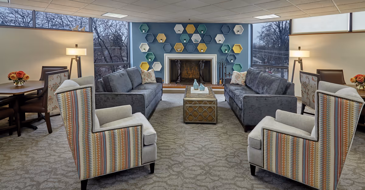 A cozy senior living room with two gray sofas facing each other, two patterned armchairs, a wooden coffee table in the center, and a fireplace on the far wall decorated with colorful hexagonal shelves. Large windows on both sides let in natural light, and there are two tables with chairs and lamps on either side of the room.