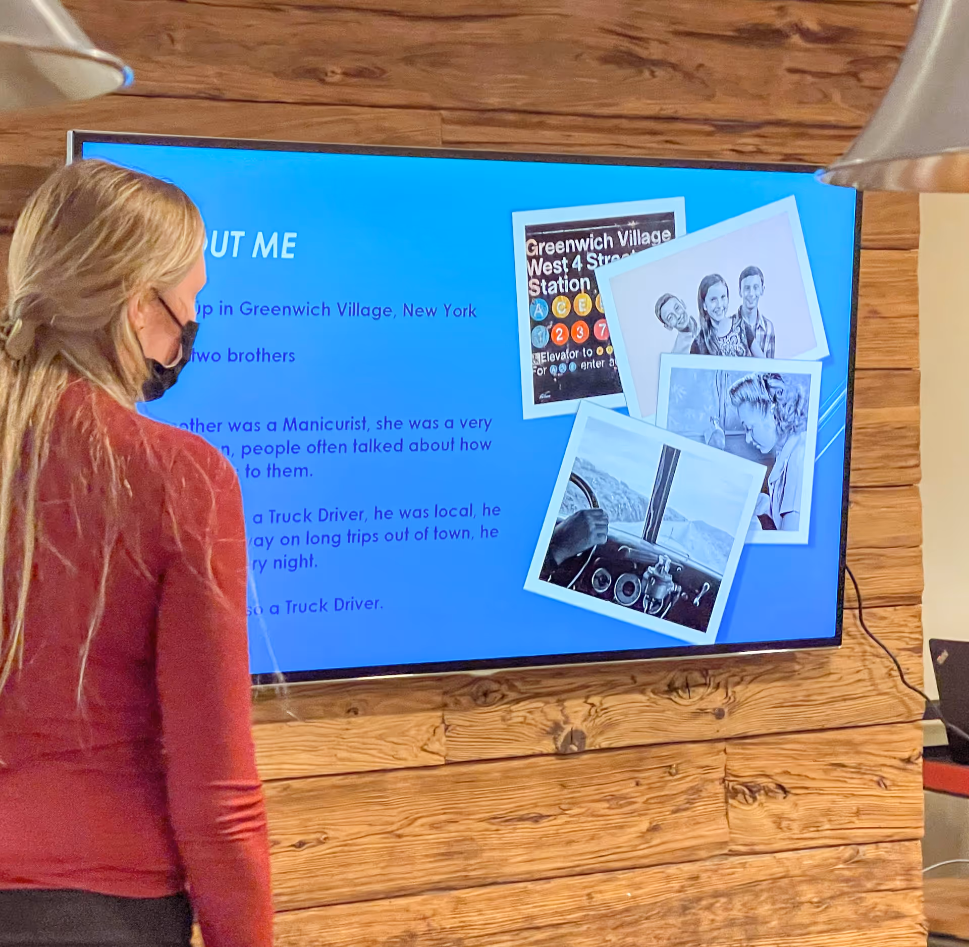 A woman wearing a face mask and a red long-sleeve shirt is standing and looking at a large wall-mounted screen displaying a blue slide titled 'ABOUT ME' with text and several photos, including a subway station sign and black-and-white portraits. The background wall is made of wooden planks.