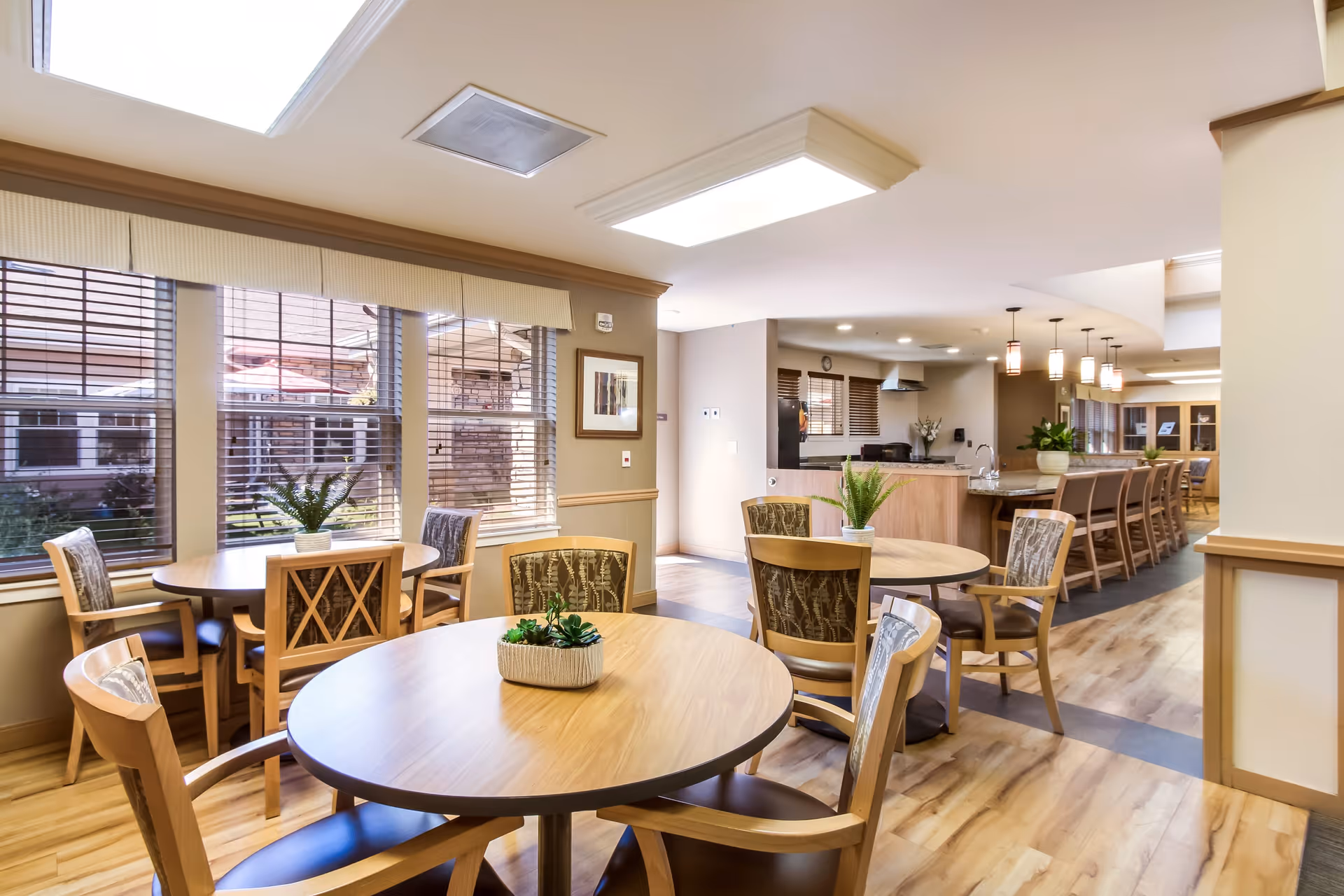 Bright and spacious dining area in Chelsea Place with round wooden tables and chairs, large windows with blinds letting in natural light, and a kitchen area with a long counter and bar stools in the background.