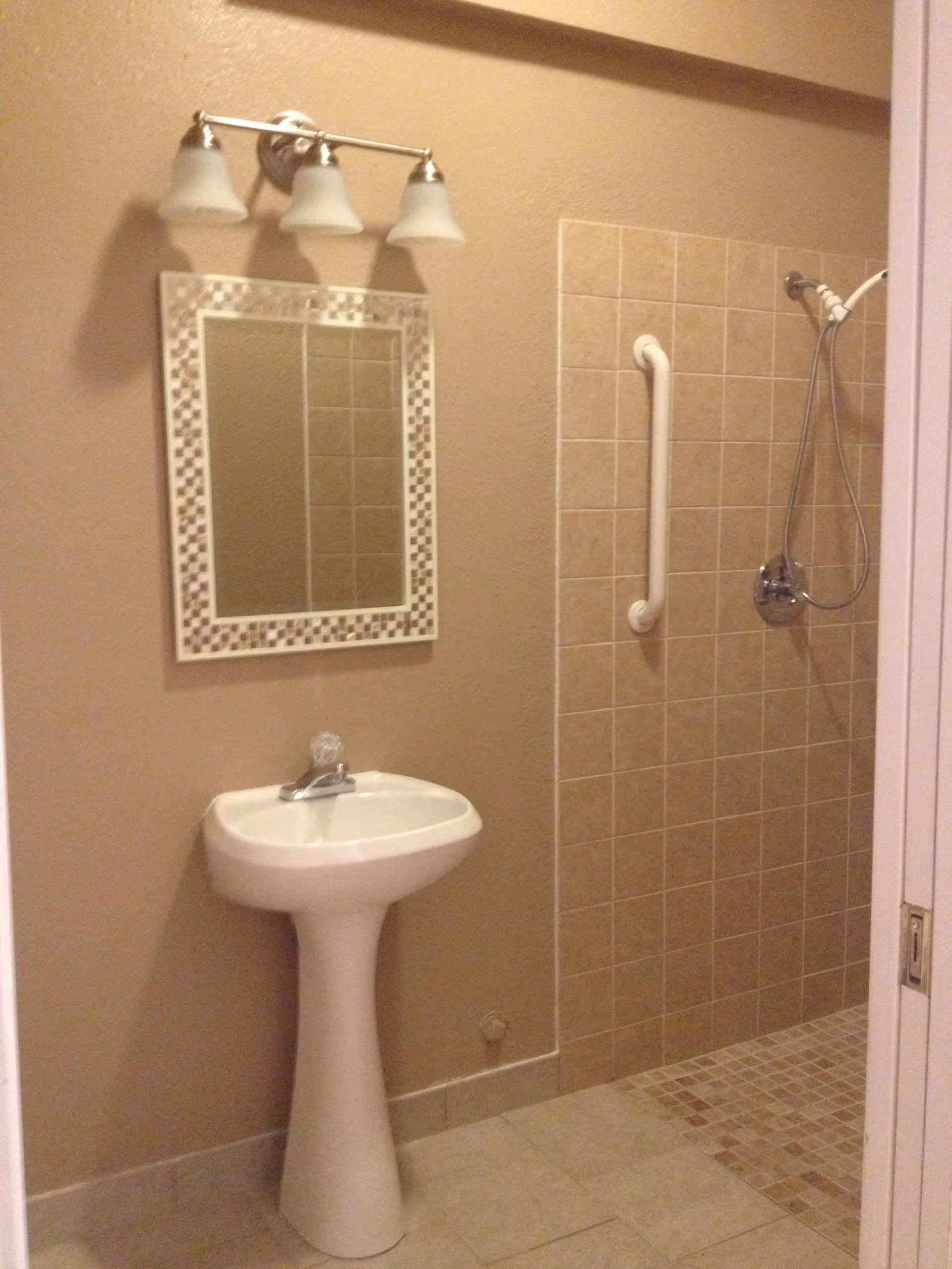 A bathroom with beige walls and tiled floor featuring a white pedestal sink with a single faucet, a rectangular mirror with a decorative mosaic frame above the sink, a three-light fixture above the mirror, and a tiled walk-in shower with a white grab bar and a handheld showerhead.