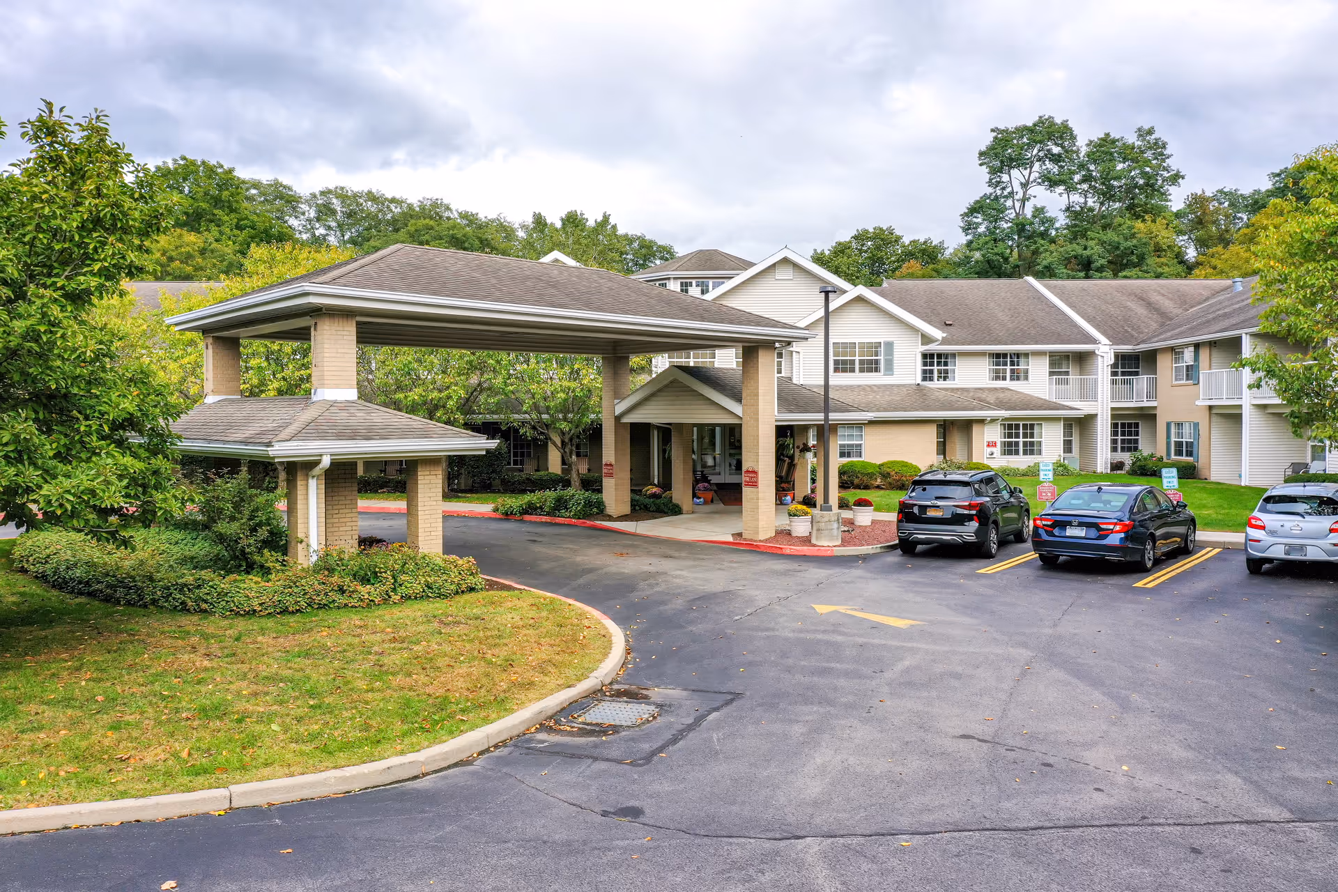 Exterior view of Solstice Senior Living at Fairport showing the main entrance with a covered drop-off area, surrounded by greenery and several parked cars in the parking lot.